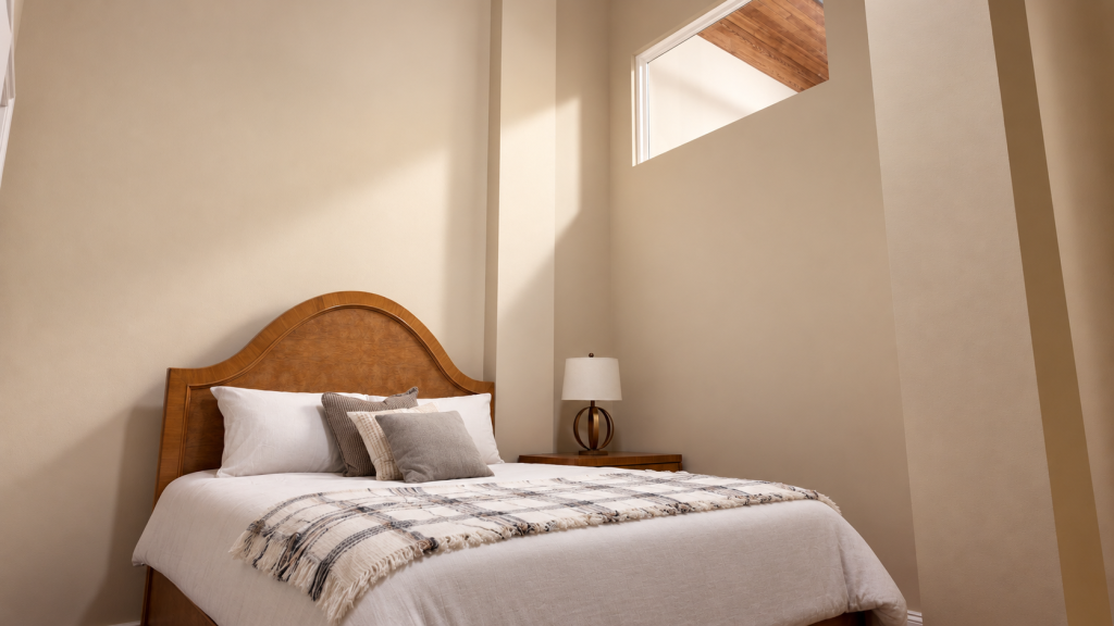 Cozy bedroom with a wood headboard bed, plaid throw, nightstand lamp, and a small high window